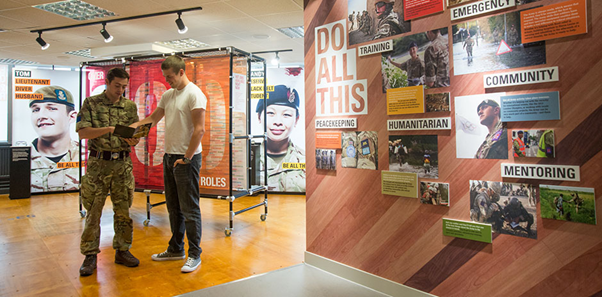 Young man in white t shirt and black trousers talking to an army officer in army outfit at a career fair with pop up boards either side featuring recruitment posters