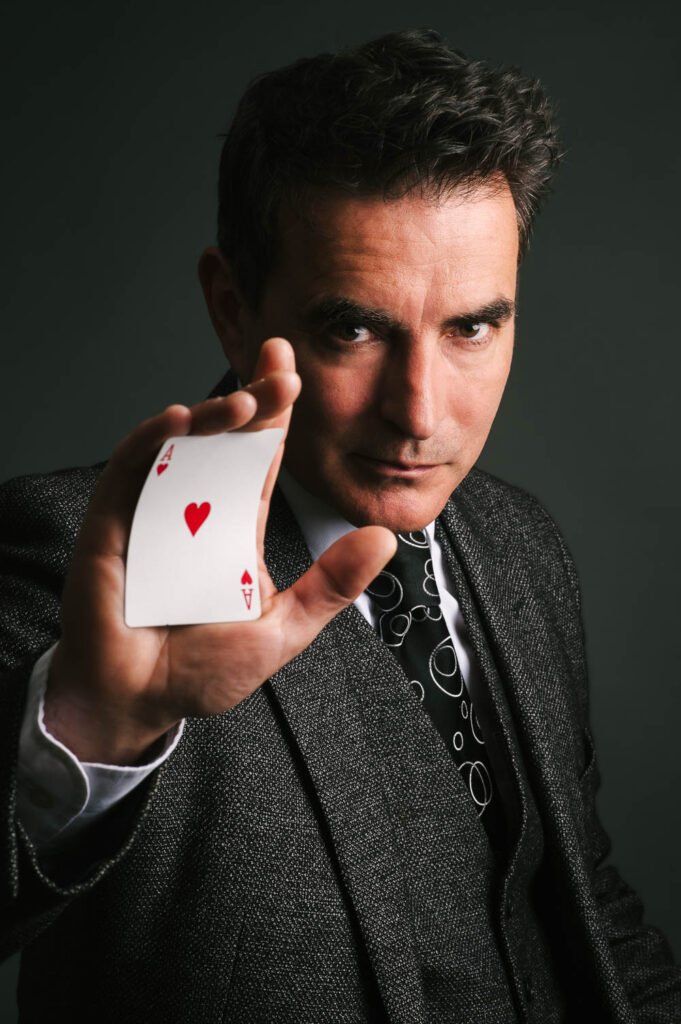 A magician with dark hair in a grey suit presenting the Ace of Hearts. 