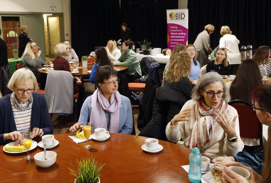 Four women at a table talking over breakfast, with groups of women at tables in the background. They are at an International Women's Day networking breakfast.