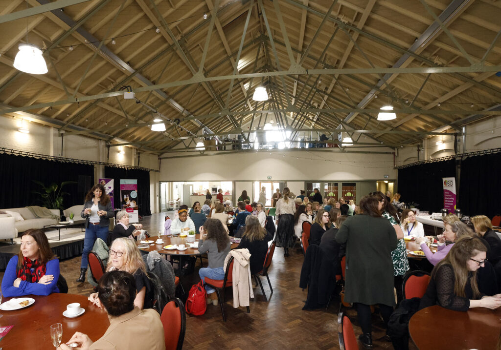 A community venue full of women at a networking event. They are sitting at tables talking to each other, with some walking around.
