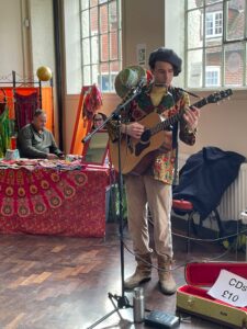 A musician in colourful, patterned clothing and a black beret plays an acoustic guitar sat Westgate Hall Market. Behind him, a vendor sits at a decorated stall selling items. A CD case marked CDs £10 is on the floor.