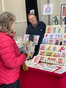 A man smiles behind a display of colourful greeting cards at a market stall, engaging with a customer in a red jacket who is browsing the cards. A sign above offers a buy four, get one free deal.