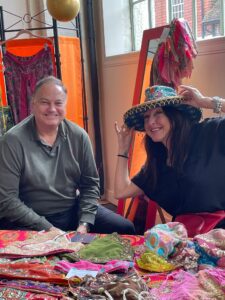 A man and a woman sit at a colourful Westgate Hall Market stall filled with vibrant fabrics and accessories. The woman is smiling and playfully tries on a decorative hat, while the man sits beside her, also smiling.