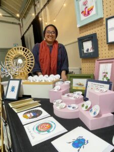A smiling person wearing glasses, a red scarf, and a dark top stands behind a table at Westgate Hall Market displaying colourful art prints and pin badges featuring insects and other designs at an indoor market stall. Framed artwork hangs on the wall behind them.