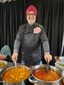 A smiling man in a red turban and black chef coat stands behind large pots of Indian curry, serving food at a stall with a Karara logo on his chest. Black curtains form the background. He is at Westgate Hall Market.