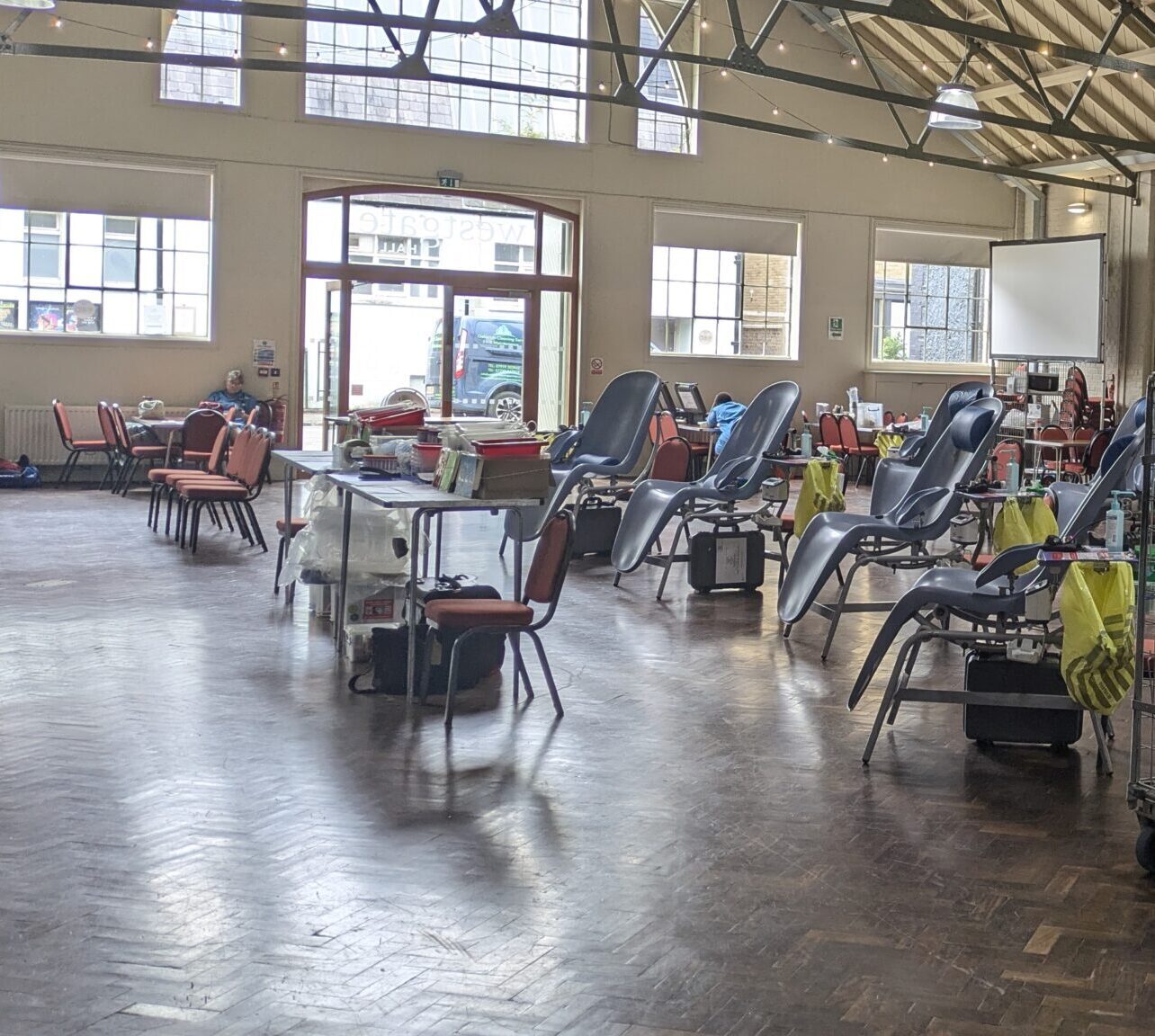 A spacious hall with rows of red chairs, several reclining donor chairs, metal carts, and medical equipment, set up for a blood donation event under high, exposed beam ceilings and large windows.